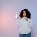 Mulher aponta para a tela em branco de um smartphone, em uma composição que remete ao uso de aplicativos de saúde e à publicidade digital.