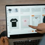 Pessoa aponta para opções de camisetas exibidas em uma loja virtual na tela de um laptop