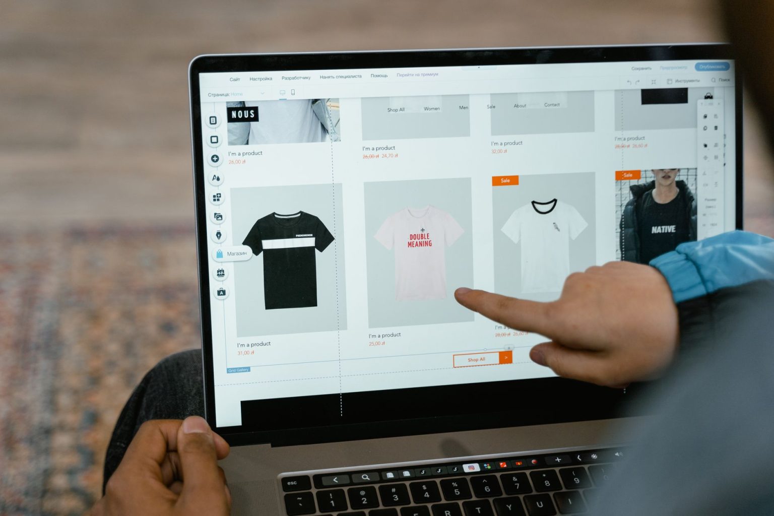 Pessoa aponta para opções de camisetas exibidas em uma loja virtual na tela de um laptop