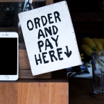 Celular ao lado de uma placa com a mensagem “Peça e pague aqui”, representando pedidos e pagamentos digitais.