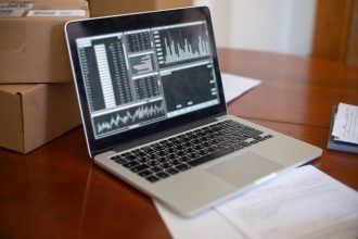 Laptop exibindo gráficos e métricas sobre uma mesa, ao lado de caixas de papelão, em um ambiente de pequeno negócio.