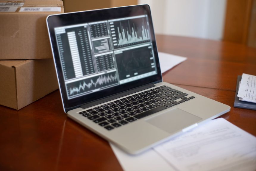 Laptop exibindo gráficos e métricas sobre uma mesa, ao lado de caixas de papelão, em um ambiente de pequeno negócio.