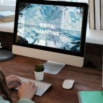 Profissional trabalha em um projeto de marketing digital em um escritório doméstico, usando computador e anotações.