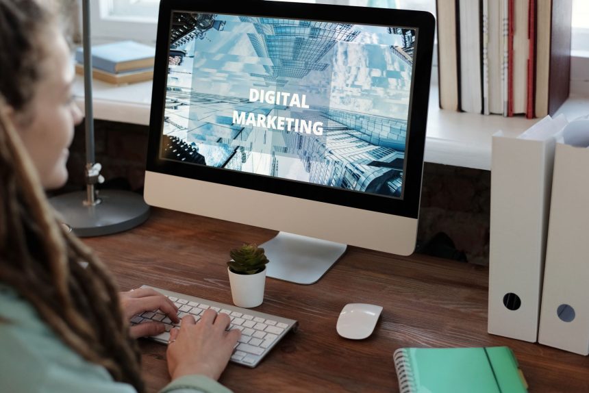 Profissional trabalha em um projeto de marketing digital em um escritório doméstico, usando computador e anotações.