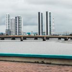 Vista do skyline de Recife, com edifícios e o rio observados a partir de uma ponte sob céu nublado.