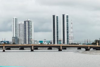 Vista do skyline de Recife, com edifícios e o rio observados a partir de uma ponte sob céu nublado.