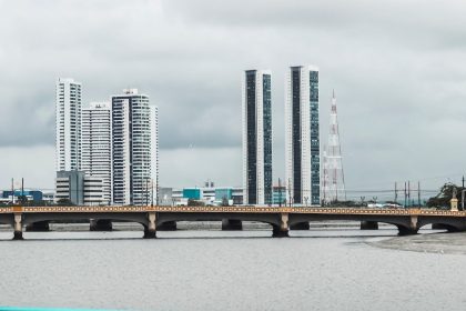 Vista do skyline de Recife, com edifícios e o rio observados a partir de uma ponte sob céu nublado.