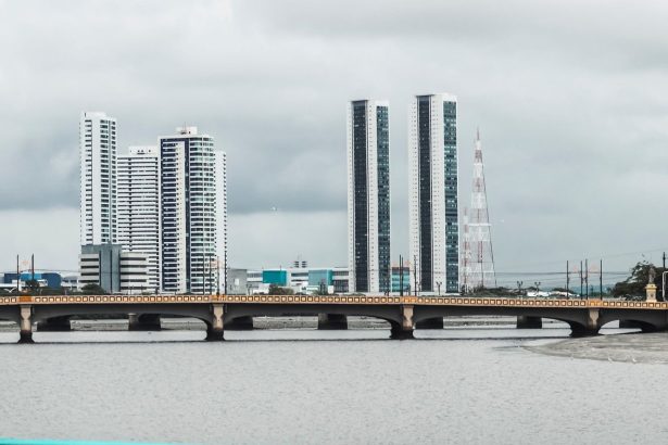 Vista do skyline de Recife, com edifícios e o rio observados a partir de uma ponte sob céu nublado.
