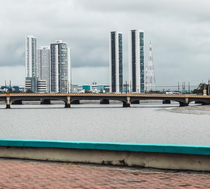 Vista do skyline de Recife, com edifícios e o rio observados a partir de uma ponte sob céu nublado.