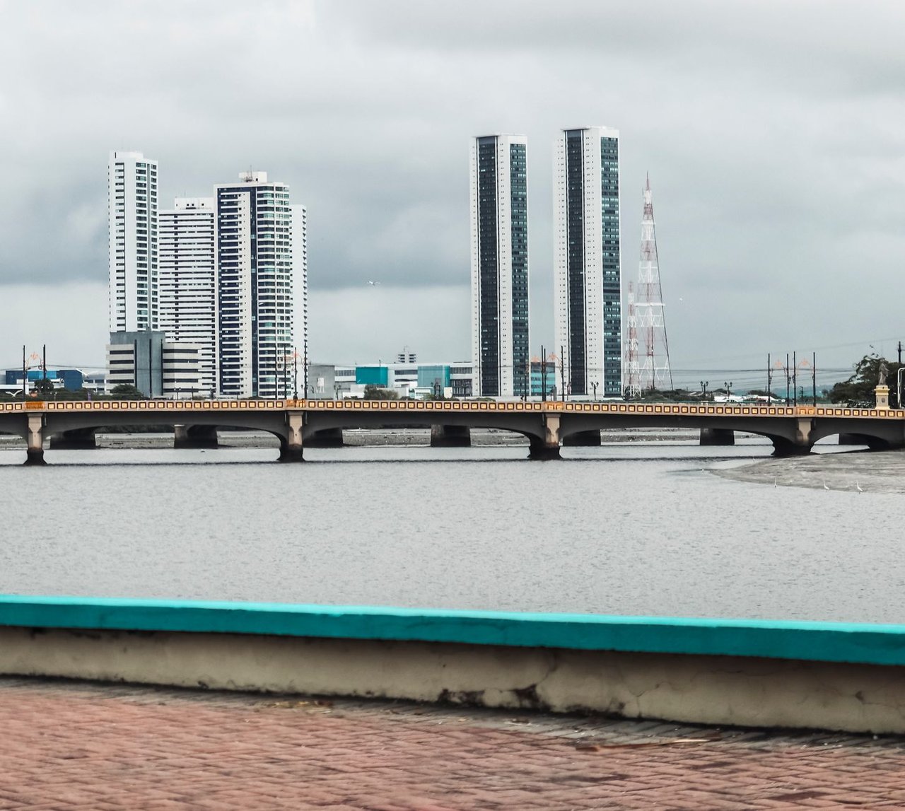 Vista do skyline de Recife, com edifícios e o rio observados a partir de uma ponte sob céu nublado.