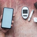 Mão segurando um smartphone ao lado de um glicosímetro e materiais médicos sobre uma superfície de madeira.