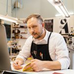 Empreendedor trabalha concentrado diante de um computador em uma gráfica movimentada.